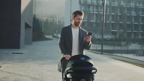 Father Using Smartphone During Morning Walk Young Father in Casual Wear Pushes Stroller Through City