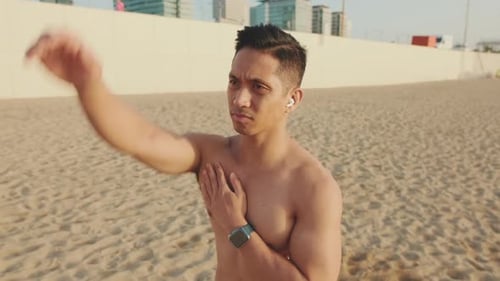Young athletic man standing on the beach stretching his arm muscles before training