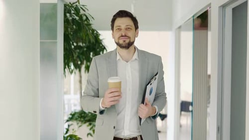 Businessman Walking Corridor in Office Holding Coffee and Tablet with Papers