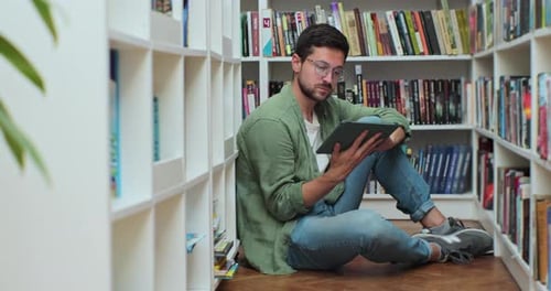 Caucasian Man Sitting on the Floor Near Shelves with Book in the Library Uses Digital Tablet