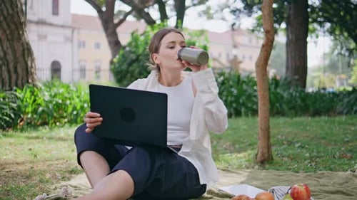 Online-Student studiert Park mit Laptop bei Picknickdecke Frau trinkt