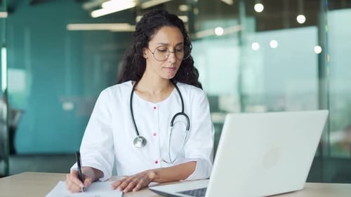 Doctor Writes Notes While Using Laptop in Office