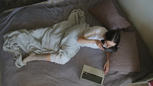 Woman Lies in Bed Typing on Laptop Wearing Headphones