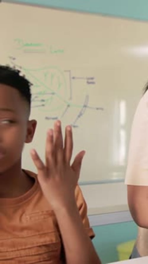 Vertical video: Listening youth in orange tee turning to adult talking about board at desk in class