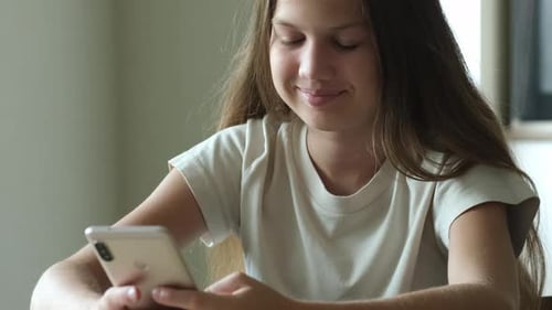 Smiling Kid Girl with Phone Sitting at Table Indoors Typing Text Chatting