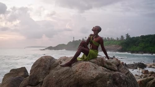 Una persona negra queer posa con un vestido abierto, joyas hechas a mano, sentada en piedra con un pintoresco acantilado marino