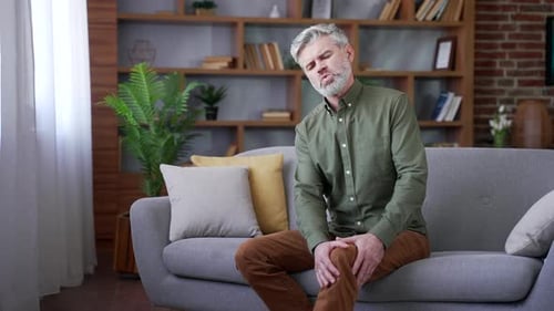 Mature Man Massages Aching Knee on Couch