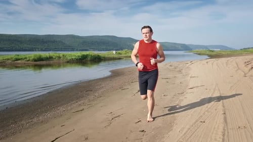 Un hombre corre en la playa. Joven deportista corre descalzo por una costa arenosa a lo largo del río. El clip tiene un plan