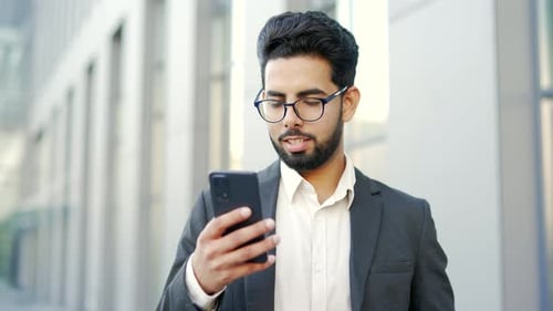 Smiling young businessman in formal suit is using a smartphone while standing on street near