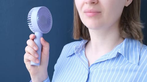 Female hand holding mini fan on blue background close-up.