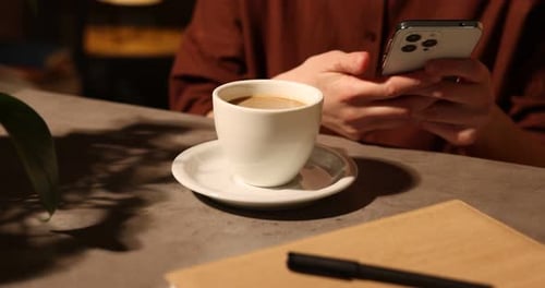 Woman using smartphone with cup of delicious coffee at grey textured table in cafe, closeup