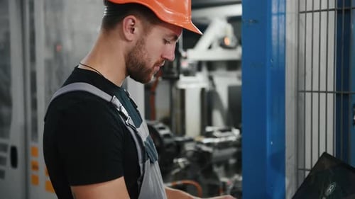 Young worker in uniform stands in the factory near automatic mechanism and uses laptop