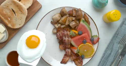 Adding A Fried Egg With A Spatula To A Breakfast Plate With Fruit, Bacon And Potatoes, Ready To S...