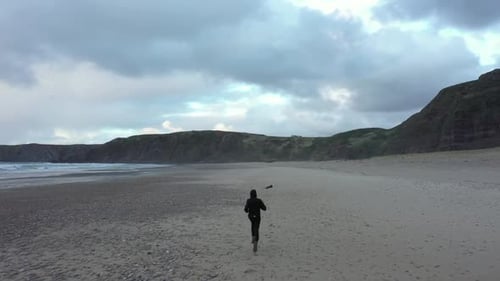 Tracking rearview aerial, man running on the sandy beach surrounded by cliffs