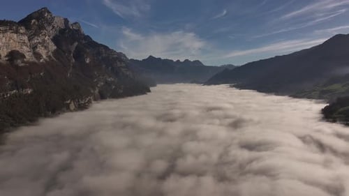 A dense fog covering a valley surrounded by rugged mountain peaks under blue sky, aerial view