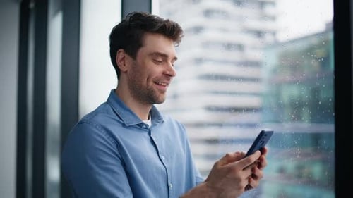 Smiling Businessman Reading Cellphone Message Standing By Office Window Closeup