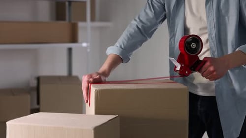 Man using dispenser with adhesive tape to seal cardboard box indoors, closeup