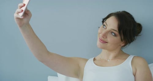 Smiling Woman Taking Selfie with Cell Phone in Bed