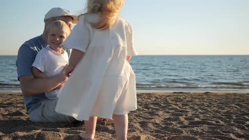 A Little Boy and Girls Resort to Their Dad and Sit on His Lap on the Beach