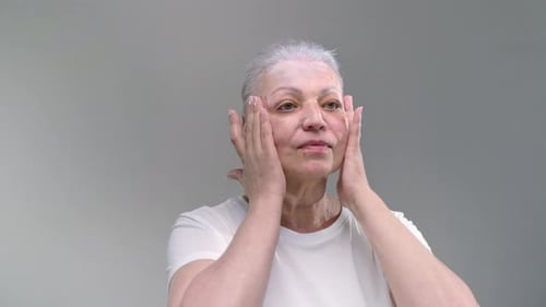 Positive Old Woman Massages Forehead and Cheeks Spreading Moisturizing Lotion on Light Background
