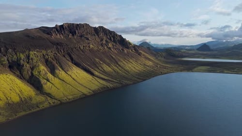 Drone pulls back over tranquil lake and green hills, revealing vast volcanic terrain