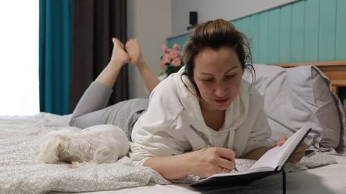 Junge Frau schreibt in den Abendstunden Tagebuch auf dem Bett im gemütlichen Schlafzimmer