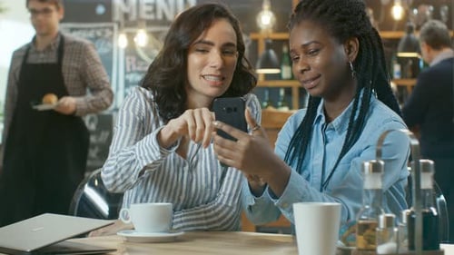 Two Beautiful Girlfriends Sitting in the Coffee Shop and Chatting, One Shows Pictures on Her Smartp
