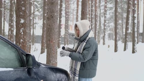 in the winter forest, a woman pours a hot drink from a thermos while standing near the machine