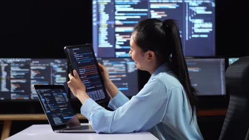 Side View Of Asian Female Programmer Looking At Database On Tablet While Writing Code By A Laptop