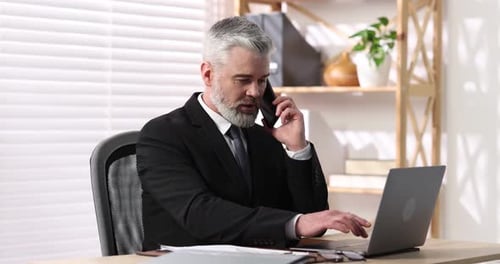 Emotional businessman in suit talking by smartphone and working with laptop at table in office