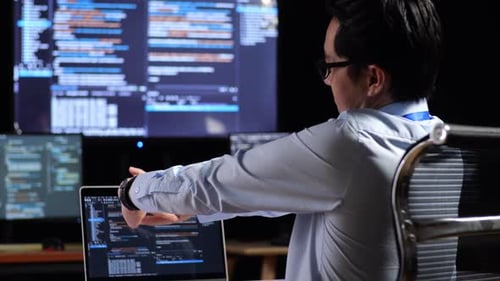 Programmer Stretching While Writing Code By A Laptop Using Multiple Monitors Showing Database