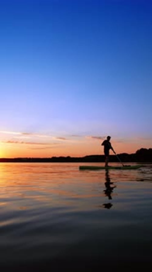 Sportsman practices paddle boarding on the river at sunset.