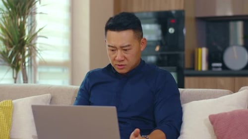 Sad Serious Asian Man Sitting on Sofa Use Laptop Computer Work at Home Network Work Unhappy Guy