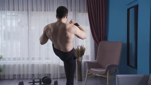 Young athlete doing kickboxing against the air at home with his back turned.