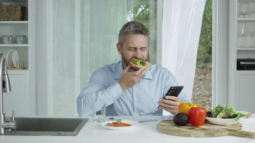 Man Eating Snack While Using Smartphone in Kitchen