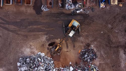 Vista desde arriba de un depósito de chatarra de metal con una excavadora amarilla que mueve la chatarra entre grandes pilas de