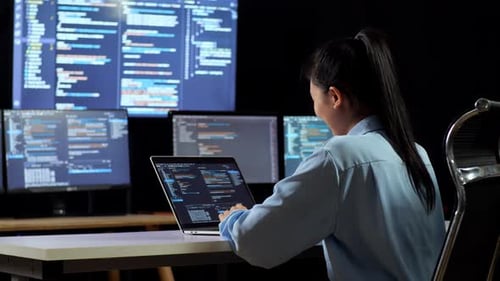 Back View Of Asian Female Programmer Writing Code By A Laptop Using Multiple Monitors