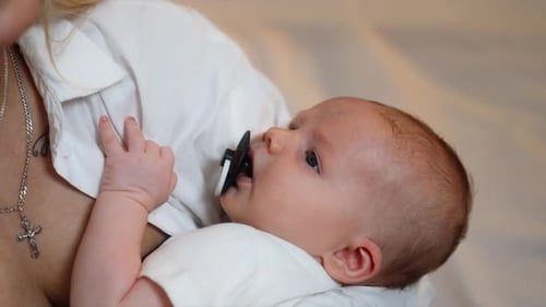 Tiny newborn is waved in mom’s hands. Baby boy suckling a pacifier and looking at mom. Close up.