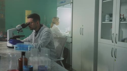 Side View of Young Caucasian Man in Lab White Coat Sitting at Workplace and Looking Into Microscope
