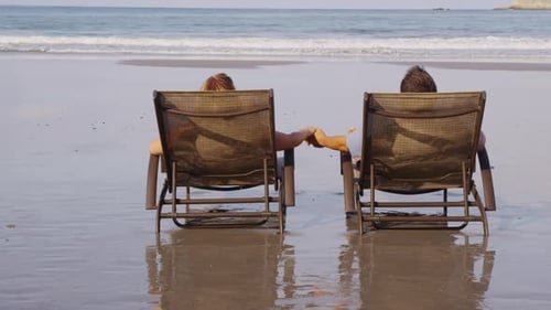 Couple Sitting In Chairs At Beach Holding Hands