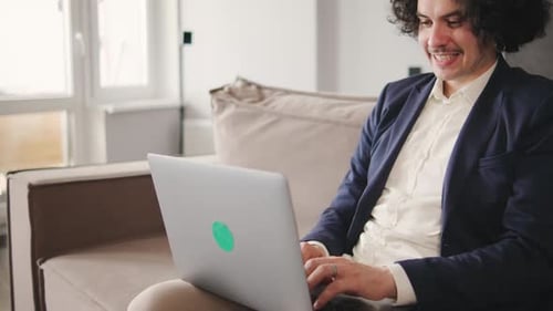 Curly Focused Business Man in a Jacket and Pajama Pants Working on Laptop Sitting at Home Office