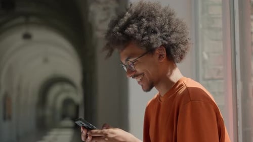 African American Male Student Smiling Using Mobile Phone Scrolling in University Corridor Happy