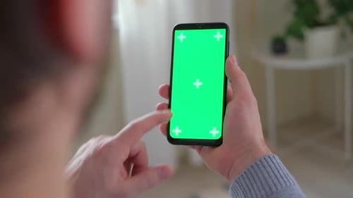 Young Man Sitting at Home Holding Smartphone Green Mockup Screen in Hand
