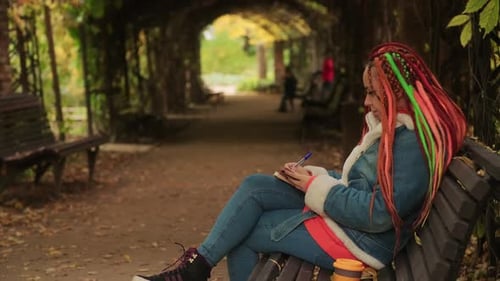 Young Woman with Dreadlocks Drawing Writing in Notebook Sitting on Bench Under Archway with