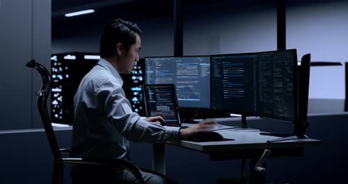 Side View Of Asian Male Programmer With A Tablet Writing Code By Desktops In The Server Room