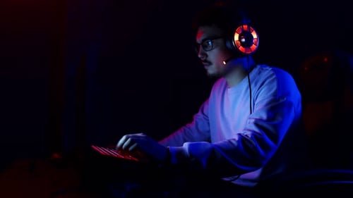 Man is Playing Online Videogames on Laptop with Headphones at Head Side View Young Man with Backlit