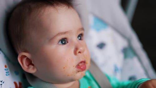 Caucasian baby dirtied with food after feeding. Little kid shoves the spoon into mouth. Close up.