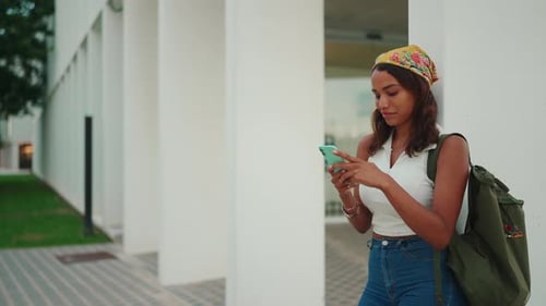 Cute tanned woman in yellow bandana with backpack on her shoulder is using mobile phone
