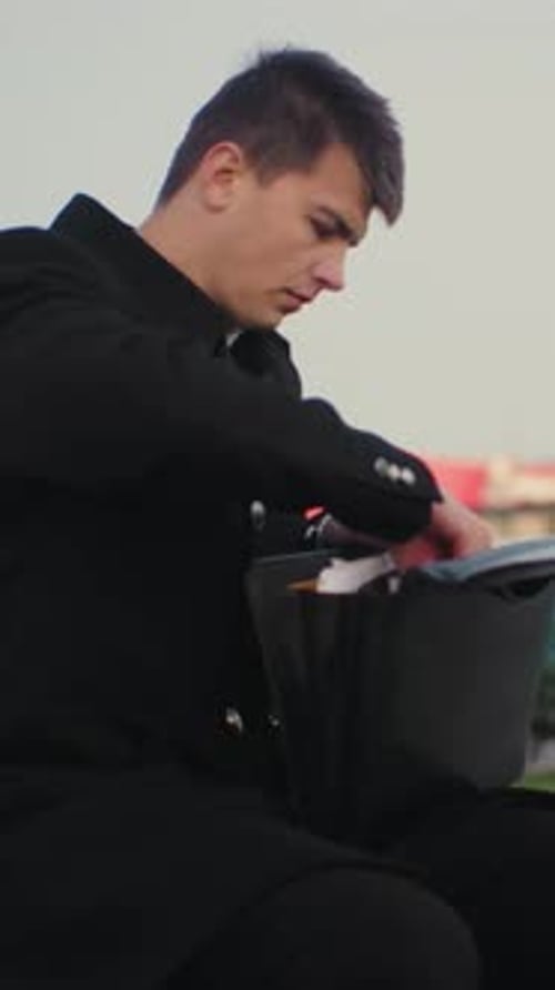 Man Seated Outdoors Going Through Briefcase Taking Out Tablet with Urban Building Background