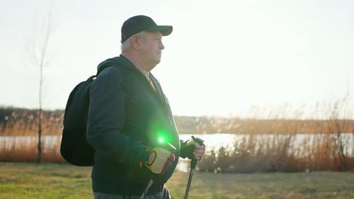 Active Handsome Elderly Man with Nordic Walking Sticks Walks Forward Through Along the Lake at